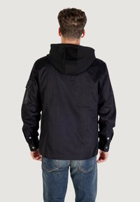 Chaqueta negra con capucha, fabricada en un tejido texturizado, con botones a presión y un bolsillo parche en la manga izquierda. Combinada con vaqueros azules.
