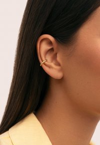 Primer plano de la oreja de una mujer con cabello largo y oscuro y un aro de oro retorcido en la parte superior de la oreja, que lleva una prenda de color amarillo pálido.