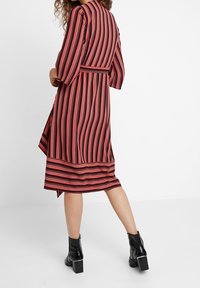 Robe longue à rayures rouges, orange et noires, avec une ceinture à la taille et des manches trois-quarts. Portée avec des bottines noires.
