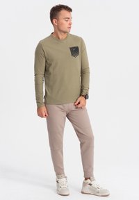 Olivgrünes Langarmshirt mit einer schwarzen grafischen Tasche, kombiniert mit hellbeigen Hosen, die Texturen und Nahtakzente aufweisen. Weiße Sneakers runden das Outfit ab.