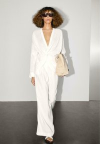 Witte wikkelblouse met een diepe V-hals, gecombineerd met loszittende witte broeken. Model draagt een geweven beige tas en heeft een paar donkere zonnebrillen op.