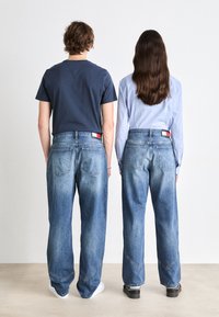 To personer står rygg mot rygg. Begge har på seg lyseblå denimjeans med en falmet finish. Jeansene har karakteristisk søm og baklommer.