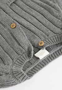 Pull gris en tricot avec texture côtelée, doté de boutons en bois et d'une étiquette cousue. Le tissu semble doux et chaud.