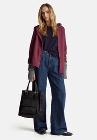 Sudadera con cremallera color burdeos, suéter azul marino, jeans de mezclilla oversized, guantes sin dedos grises, bolsa de mano negra con detalles metálicos y zapatos planos negros.