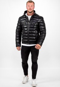 Giacca a vento nera con chiusura a zip, design trapuntato, colletto alto e due tasche con zip; indossata con jeans neri strappati e sneakers bianche.