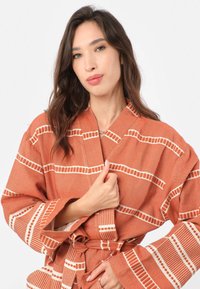 Rúžovo-hnedý plášť s bielymi pruhovanými vzormi, vyrobený z textúrovanej tkaniny. Má opasok na zapínanie a široké rukávy pre pohodlie.