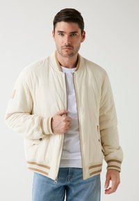 Chuněná béžová bomber bunda s žebrovanými manžetami a lemem, s odnímatelným zipem a béžovými pruhy. Nositelná přes bílý tričko.