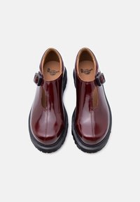 Παπούτσια από δερμάτινο βερνίκι μπορντό με σχέδιο T-strap, με ρυθμιζόμενο πόμολο και χοντρή μαύρη καουτσούκ σόλα.