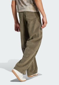 Olive green corduroy pants with wide legs, ribbed texture, and two rear pockets, paired with white sneakers and light-colored shirt.