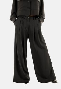 Donna che indossa pantaloni neri a vita alta e a gamba larga con pieghe, le mani nelle tasche, abbinati a un giubbotto nero cropped con bottoni e scarpe nere.