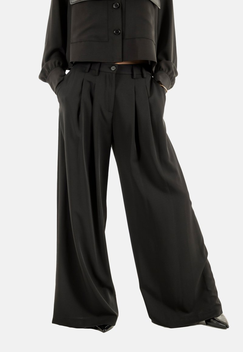 Donna che indossa pantaloni neri a vita alta e a gamba larga con pieghe, le mani nelle tasche, abbinati a un giubbotto nero cropped con bottoni e scarpe nere.