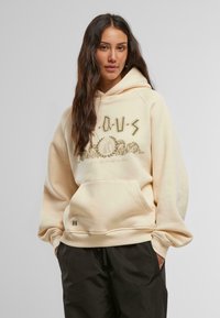 Beige, oversized kapucnis pulóver zsinóros kapucnival, elülső zsebbel és arany grafikával. Puha anyag, bordás mandzsettákkal és aljjával.