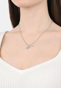 Collana d'argento con pendente a forma di chitarra, caratterizzata da una catena sottile e una texture liscia, indossata sopra un indumento bianco a scollo a V.