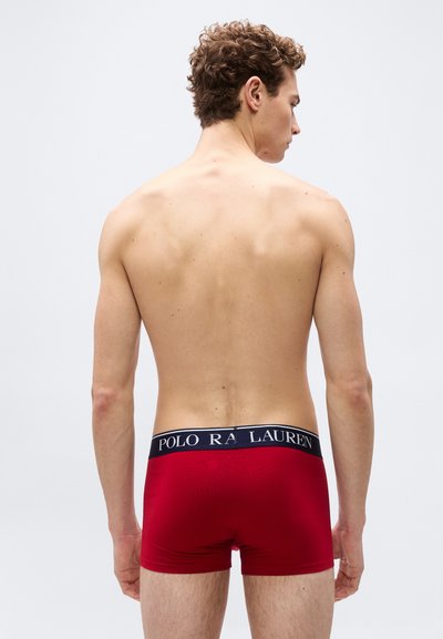 Άνδρας με σγουρά μαλλιά, όρθιος με την πλάτη στην κάμερα, φοράει κόκκινα μποξέρ με μαύρη ζώνη μέσης που γράφει "Polo Ralph Lauren."