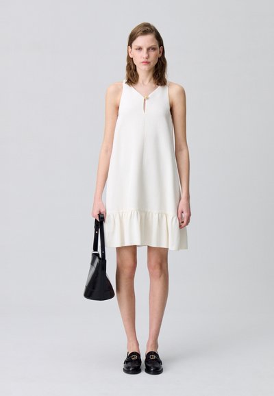 Vestido blanco sin mangas con un detalle en forma de ojal en el escote y un dobladillo con volantes. Combinado con mocasines negros y un bolso negro pequeño.