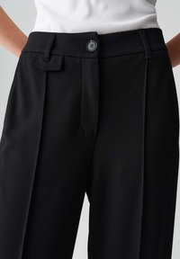 Pantaloni neri su misura con chiusura a bottone e pieghe frontali, indossati con un top senza maniche a costine bianco.