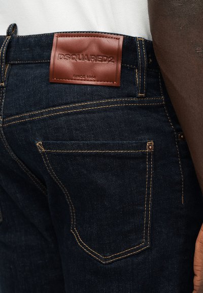 Σκούρα μπλε τζιν από τζιν ύφασμα με κίτρινη ραφή και καφέ δερμάτινη ετικέτα με την επιγραφή "DSQUARED2 SINCE 1964" στη πίσω μέση της ζώνης.