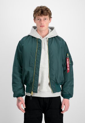 Veste bomber en nylon verte avec des poignets côtelés, fermeture éclair et une étiquette décorative rouge sur la manche. Portée par-dessus un sweat à capuche gris.