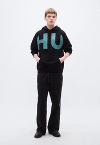 Μαύρο φούτερ με κουκούλα και μεγάλο τυρκουάζ λογότυπο "HU", συνδυασμένο με μαύρη φόρμα με μικρό τυρκουάζ λογότυπο. Λείο ύφασμα, άνετη εφαρμογή.