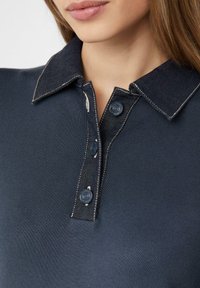 Granatowa koszulka polo z deninowym kołnierzykiem, z trzema guzikami na plastrze oraz kontrastowymi szwami. Gładka tekstura i dopasowany krój.