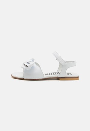 White flat sandals with an open toe, featuring a bow accent at the front, buckled ankle strap, and a flat brown sole with logo print.