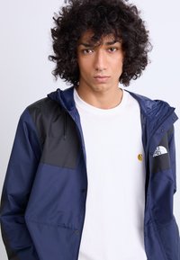 Jūras spēku un melnā krāsā, viegla, ūdensizturīga jaka ar kapuci, ar spiedpogām un logotipu uz krūtīm, virs baltas t-kreklu.