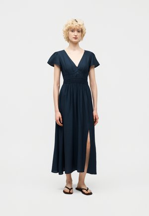 Donna con capelli biondi ricci che indossa un abito blu navy con maniche svolazzanti, scollo a V, vita aderente, spacco laterale e sandali neri su sfondo bianco.