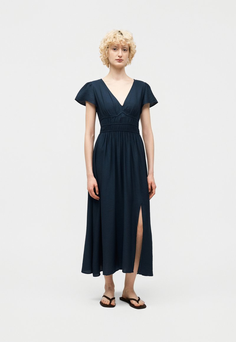 Γυναίκα με σγουρά ξανθά μαλλιά φοράει φόρεμα ναυτικό μπλε με μανίκια που φτερουγίζουν, V-λαιμόκοψη, ζωνάκι στη μέση, σκίσιμο στο πλάι και μαύρα πέδιλα σε λευκό φόντο.