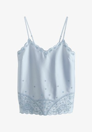 Femme portant un haut sans manches bleu clair avec broderie florale et un jean bleu foncé, debout avec le bras gauche détendu le long du corps.