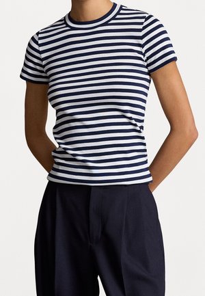 Osoba ubrana w dopasowaną koszulkę w poziome paski w kolorze granatowym i białym, z krótkim rękawem, włożoną do wysokich, granatowych spodni.