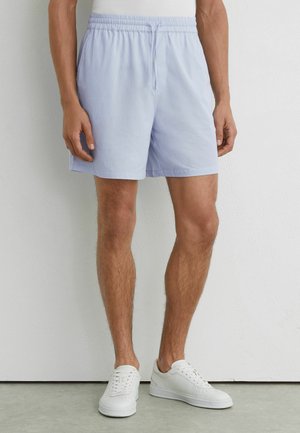 Mężczyzna ubrany w jasnoniebieskie, paskowane szorty z elastycznym pasem oraz białe trampki, stojący na tle gładkiej jasnej ściany.