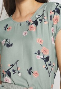 Blommönstrad kortärmad topp i mjuk grön tyg; har rosa och mörkblå blommor med rund halsringning och avslappnad passform.