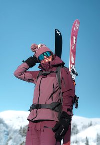 Équipement de ski comprenant une veste et un pantalon violets, des gants noirs et un bonnet rose et noir. Deux skis sont visibles, l'un rose avec un motif.