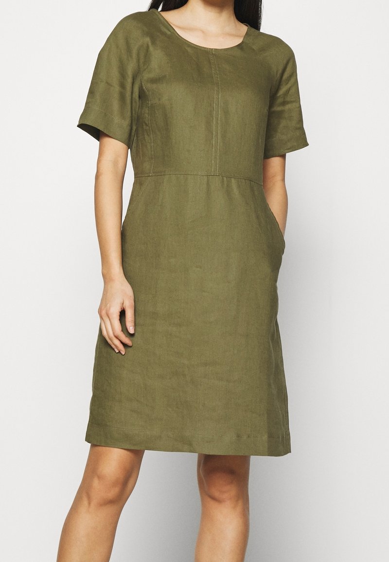 Femme portant une robe olive mi-longue avec manches courtes et poches avant, debout devant un fond uni.