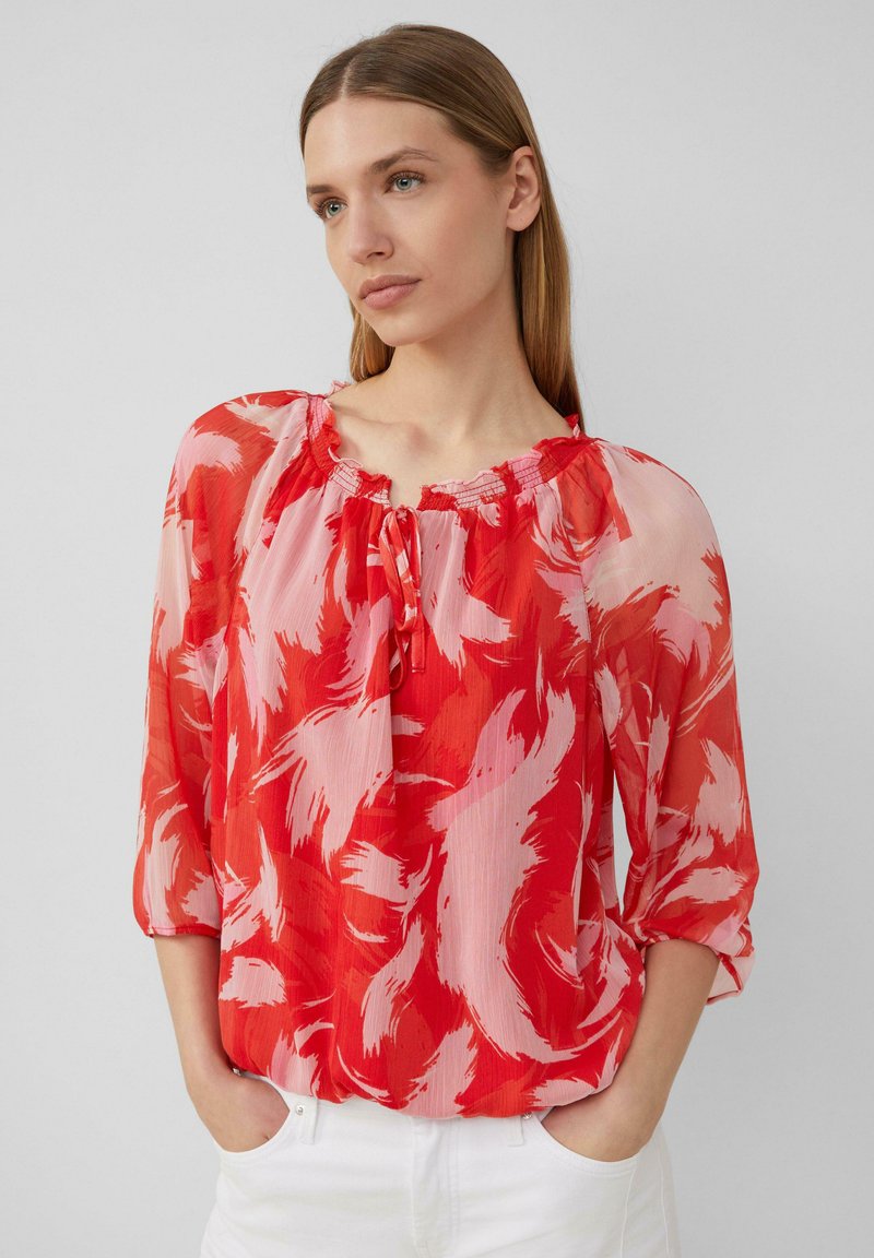 Blouse à motifs rouges et roses avec un col froncé, un détail à nouer et des manches trois-quarts, fabriquée dans un tissu léger et translucide.