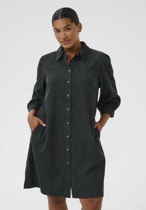 Woman with dark hair in low bun wearing dark buttoned corduroy dress with collar and three-quarter sleeves, hands in pockets, neutral background.