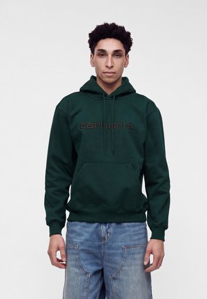 Giovane uomo con capelli ricci che indossa una felpa con cappuccio Carhartt verde scuro e jeans azzurri su uno sfondo bianco uniforme.