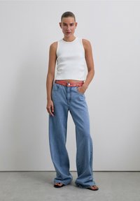 Top sin mangas de rayas blancas combinado con vaqueros holgados de un azul claro, que cuentan con una cinturilla roja con patrón. La modelo está de pie con una mano en el bolsillo.