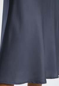 Ourlet de jupe en satin gris foncé tombant juste en dessous du genou sur une personne debout avec les jambes nues visibles.