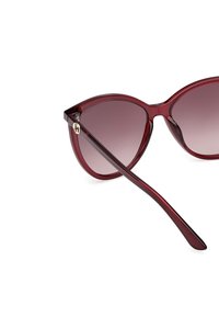 Gafas de sol de ojo de gato con montura burdeos transparente, lentes degradados y varillas delgadas; presentan un detalle de logo dorado cerca de la bisagra.