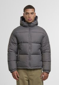 Puffjakke i grått med hette, glidelås foran og ribbestrikkede mansjetter. Designet inkluderer quiltede seksjoner for isolasjon.