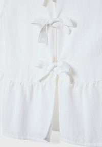Biała bezrękawnikowa bluzka z falbaniastym dołem, z przodu wiązana na wstążki z materiału oraz o gładkiej fakturze. Prosty i minimalistyczny design całości.