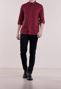 Camisa de botones a cuadros rojos y negros con cuello, mangas largas enrolladas hasta los codos, combinada con pantalones negros ajustados y zapatos negros.