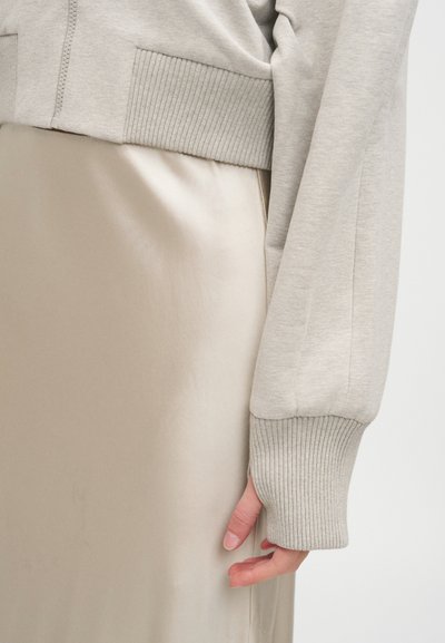 Gros plan d'une personne portant un sweat-shirt court côtelé beige clair et une jupe beige brillante, la main détendue le long du corps.