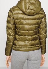 Chaqueta acolchada verde oliva con capucha, usada sobre pantalones acanalados gris claro, mostrada desde atrás en una persona de pie.