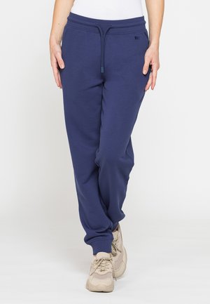 Persona che indossa pantaloni della tuta blu navy con coulisse e scarpe da ginnastica beige, in piedi con le gambe incrociate davanti a uno sfondo chiaro e uniforme.