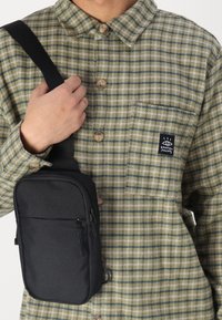 Svart tvärsöver väska i strukturerad tyg med ett zippered fack och en justerbar rem, bärs över en grön och beige rutad skjorta.