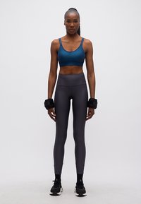 Αθλητική γυναίκα με μπλε αθλητικό σουτιέν, σκουρόχρωμο ψηλόμεσο κολάν, μαύρα βάρη αστραγάλου και μαύρα αθλητικά παπούτσια, που στέκεται μπροστά σε λευκό φόντο.