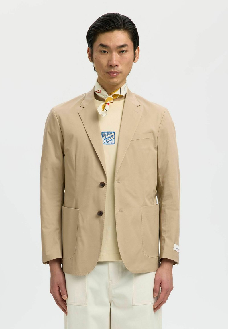 Homme portant un blazer beige sur une chemise crème avec un logo bleu, un pantalon blanc et un foulard jaune à motifs autour du cou, debout devant un fond uni.