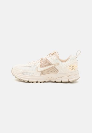 Tan and cream athletic sneakers feature mesh panels, a cushioned sole, and geometric accents on the heel. Laces integrated into the design.